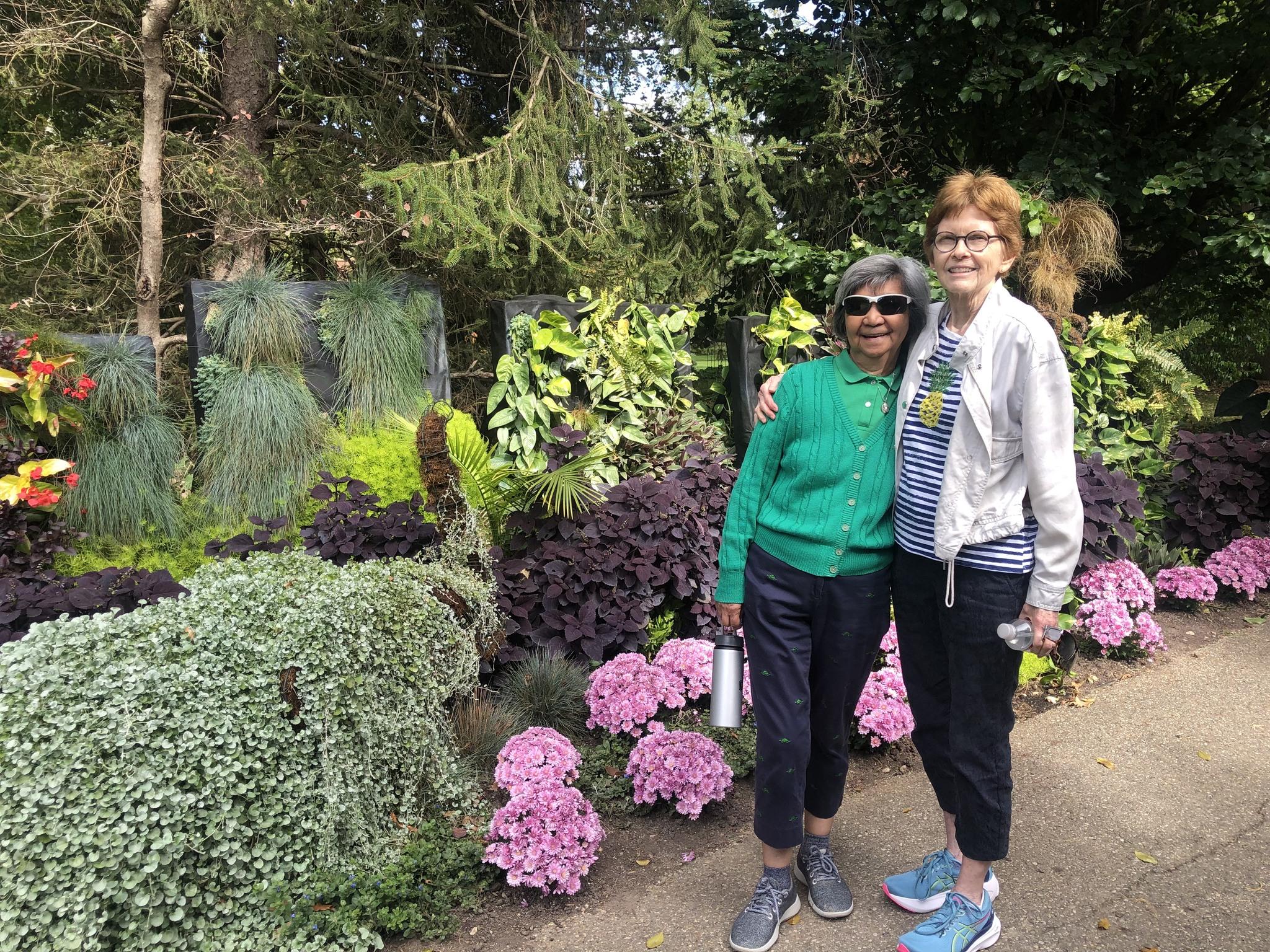 Two smiling women standing together in a lush garden with pink hydrangeas.