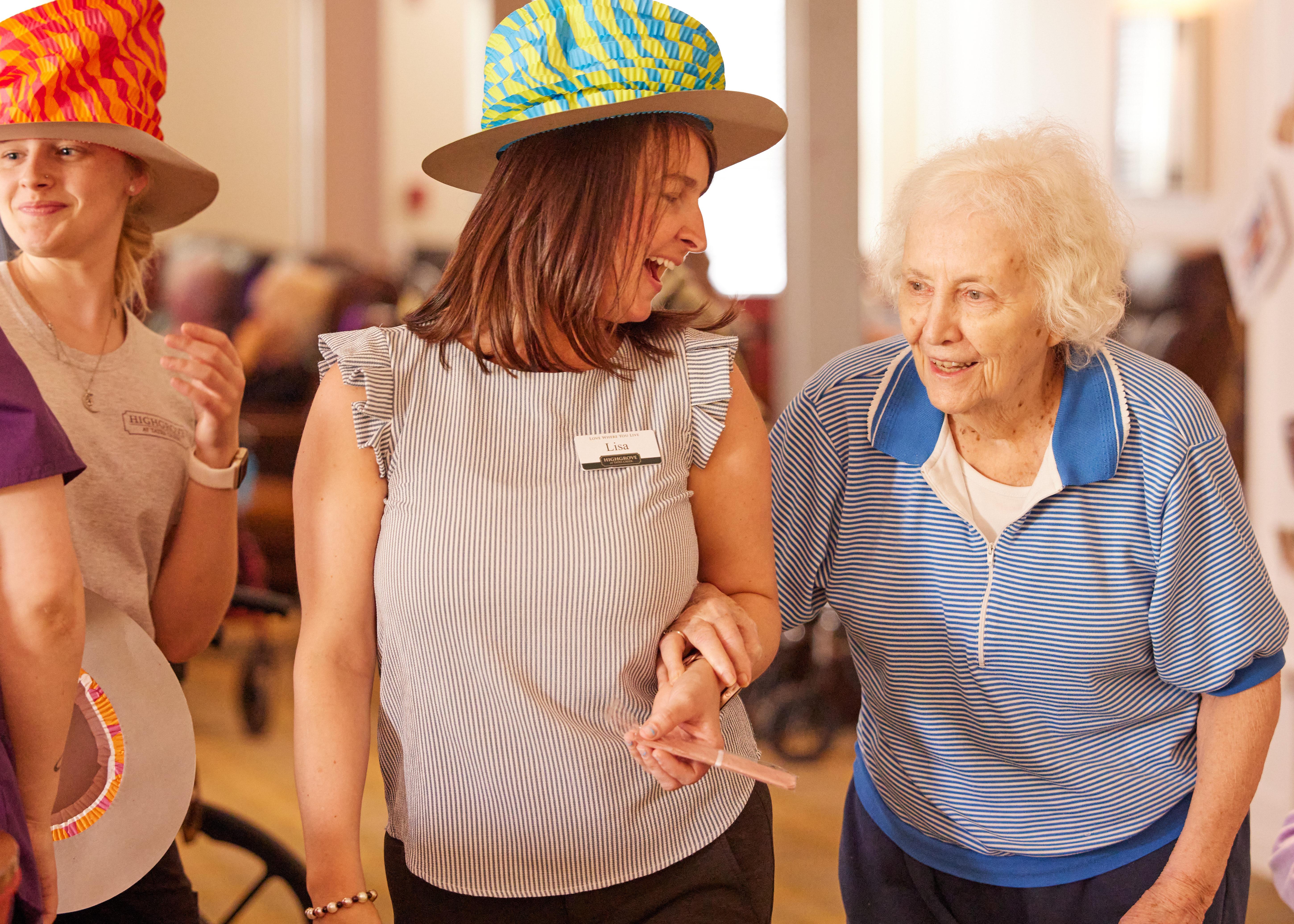A caregiver in a colorful hat dances with a smiling woman in a blue top.