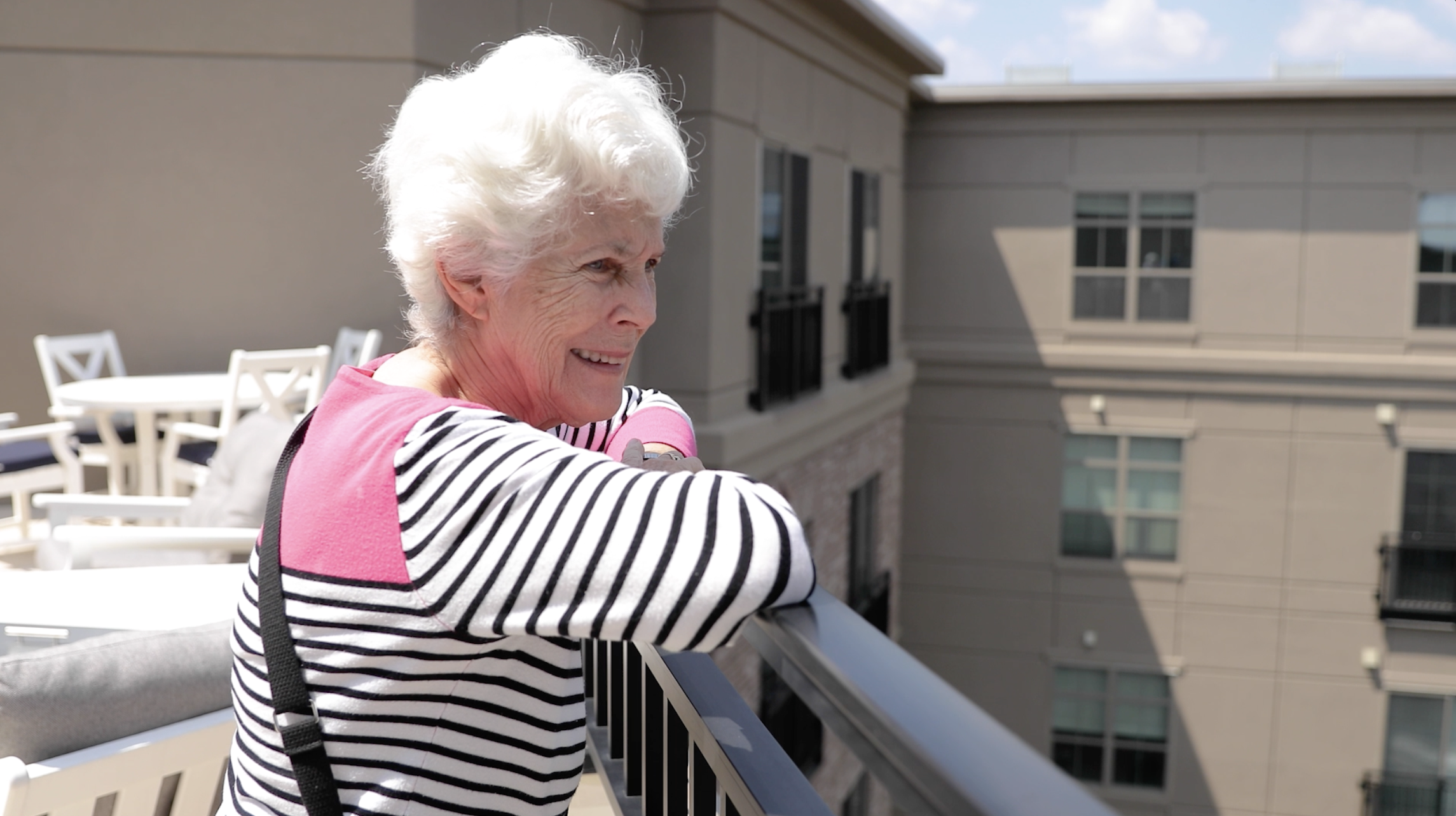 Woman with white hair smiling and looking out from a sunny balcony.