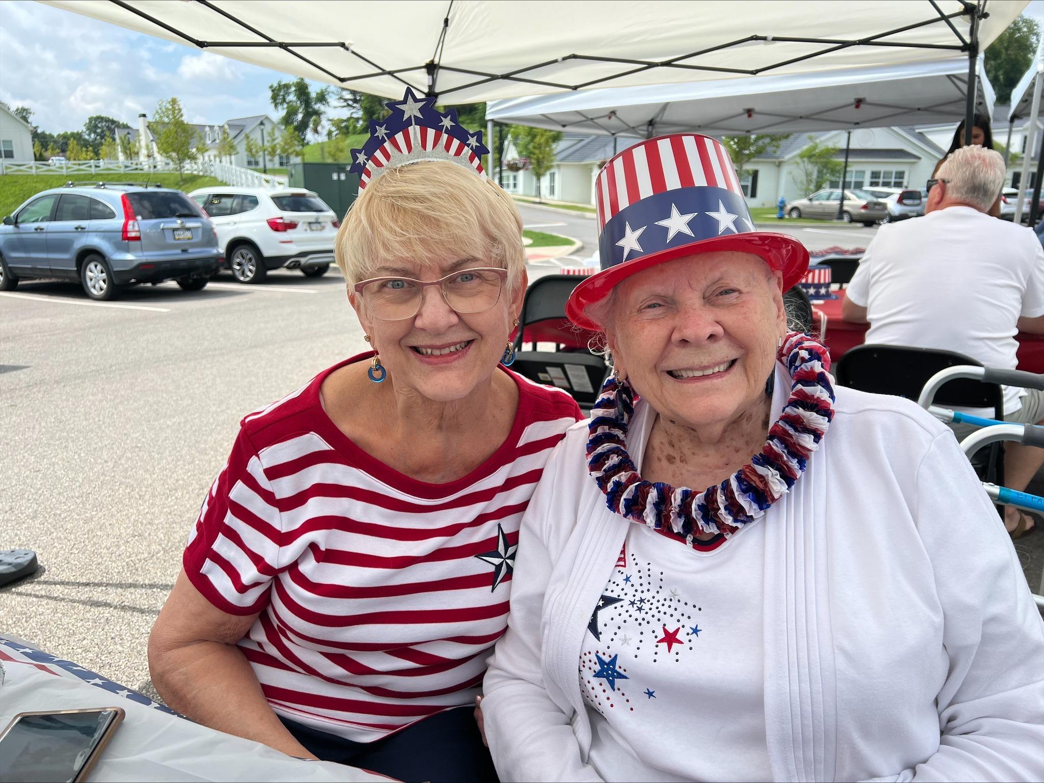 Two smiling people in patriotic attire, one wearing a stars-and-stripes hat, at an outdoor event.