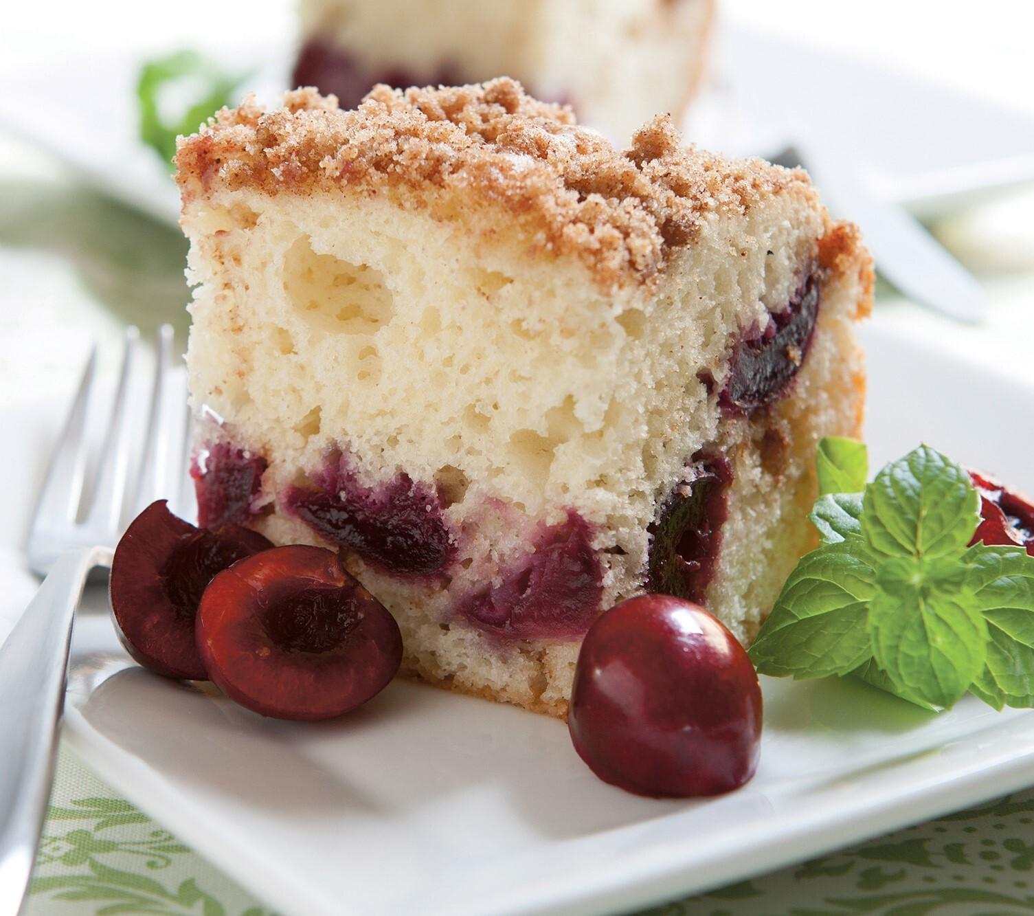 Slice of crumb-topped cherry cake on a plate with fresh cherries and mint leaves.