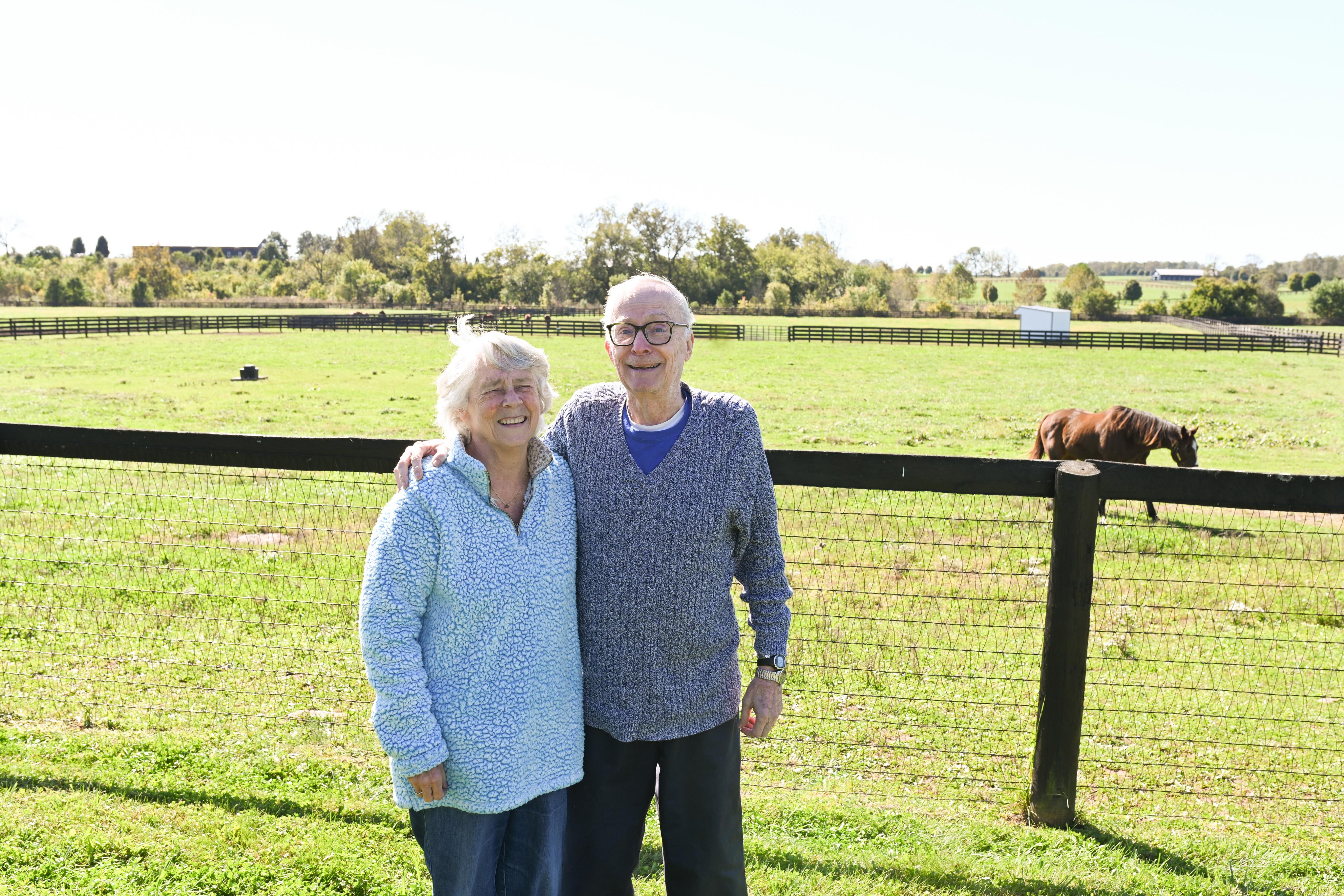 Two smiling people standing by a fence with a pastoral field and horses in the background.