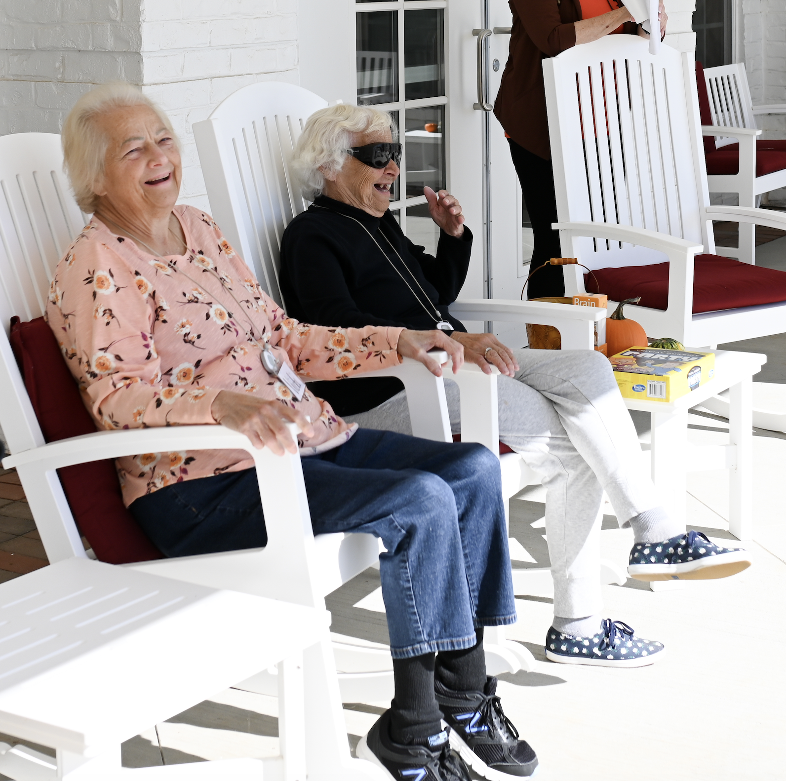 Two people relaxing in white rocking chairs on a sunny porch, smiling and chatting