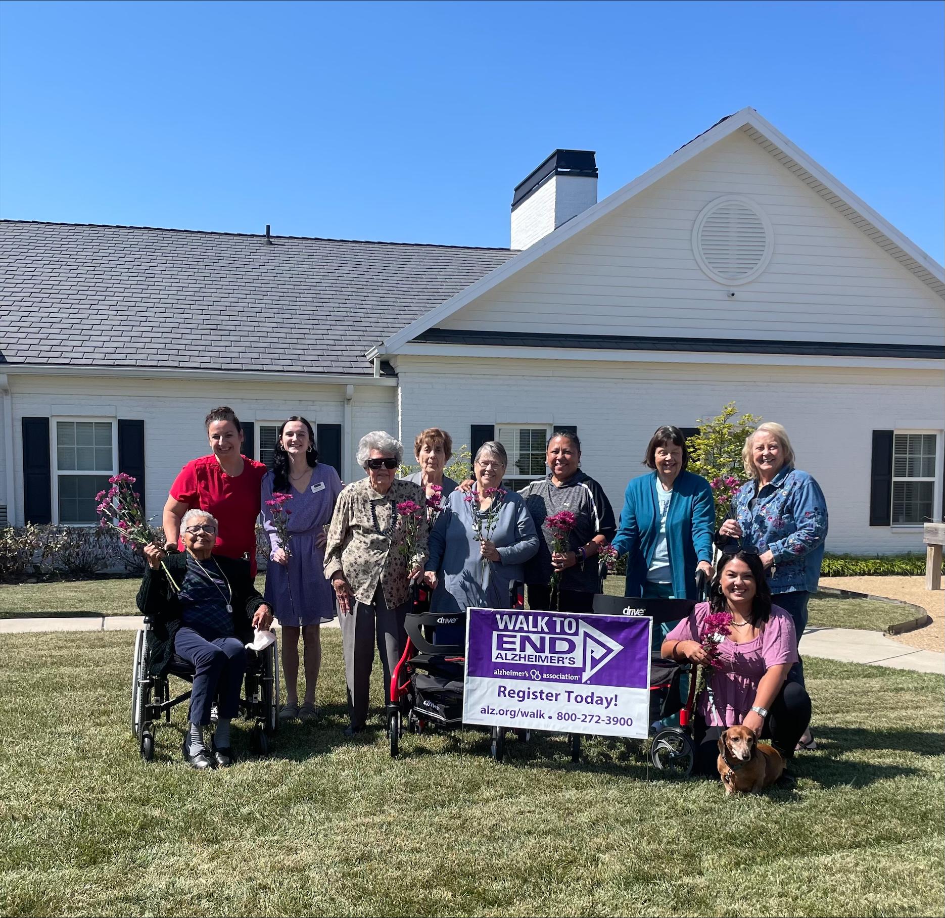 Group of people standing outdoors with a 'Walk to End Alzheimer's' sign, some holding flowers.