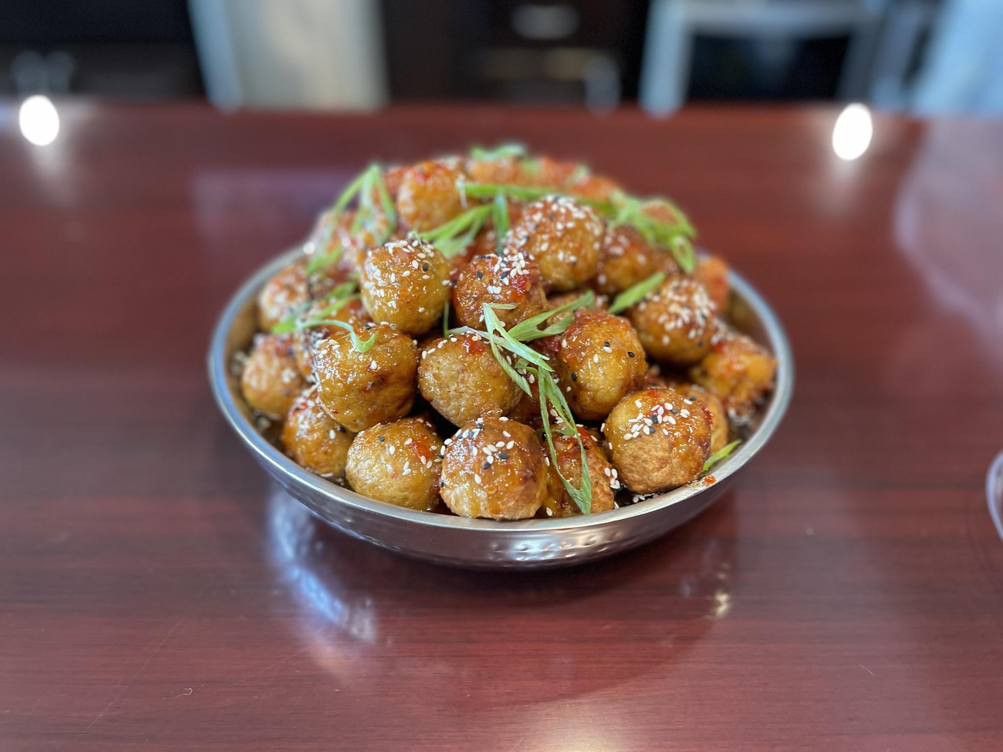 Bowl of glazed meatballs sprinkled with sesame seeds and garnished with green herbs on a table.
