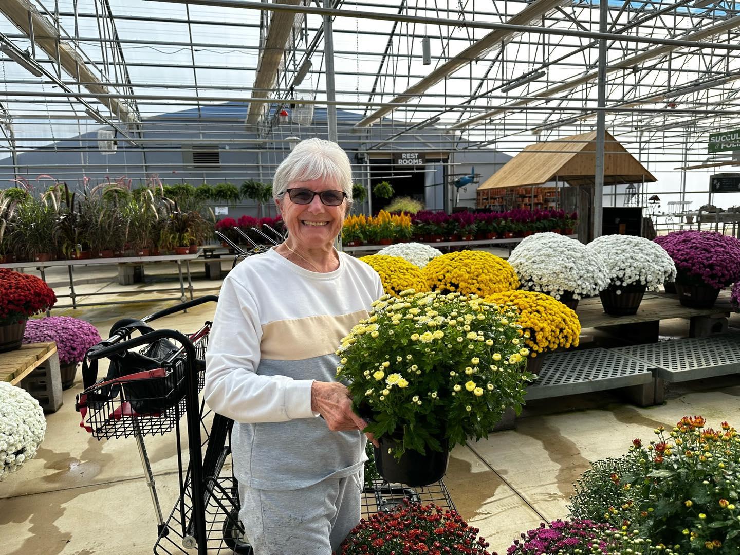 A smiling person holding a potted plant at a garden center with colorful flowers around