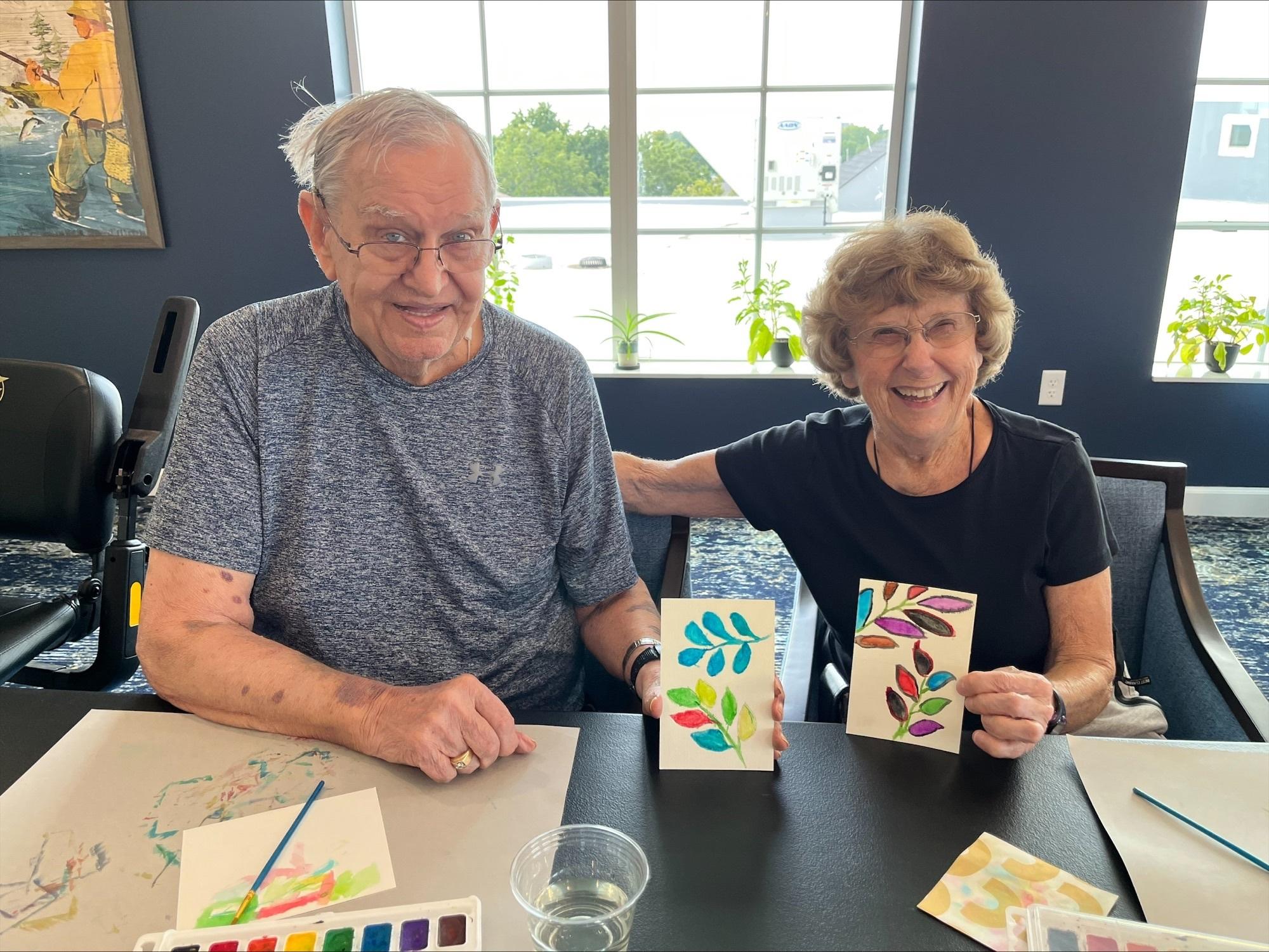 Two people smiling, displaying their painted canvases at a craft table with brushes.