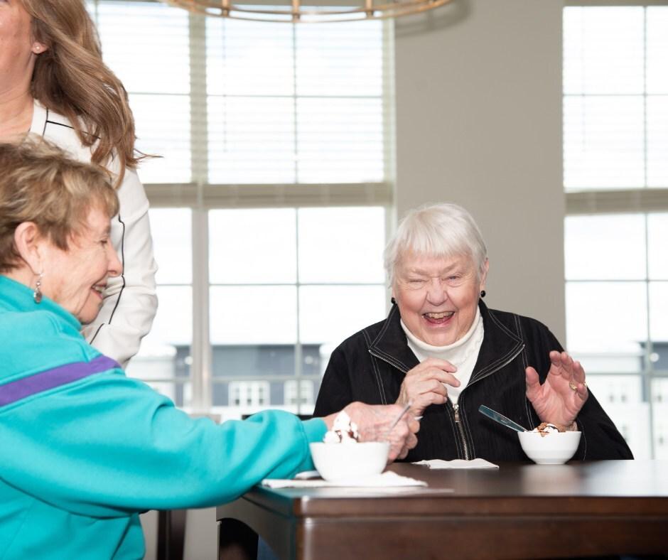 Laughing people enjoying ice cream bowls at a table with a caregiver standing by.