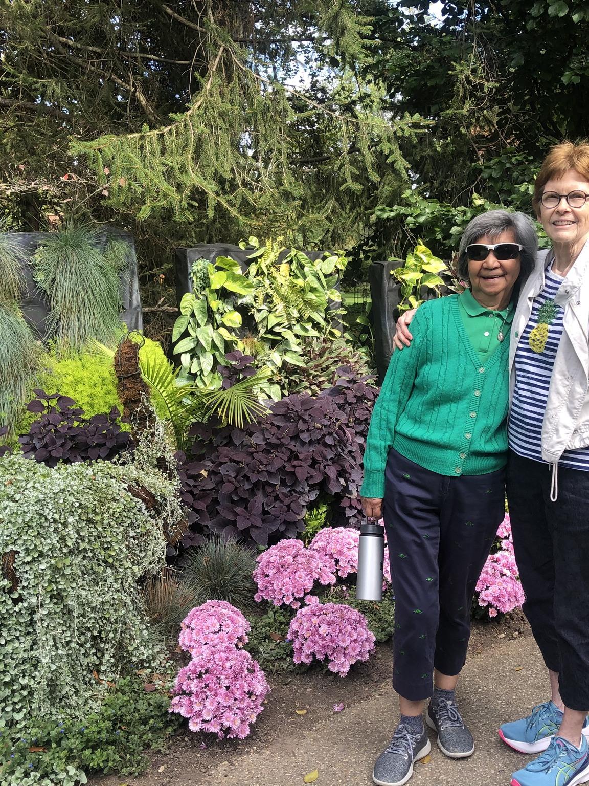 Two smiling women standing together in a lush garden with pink hydrangeas.