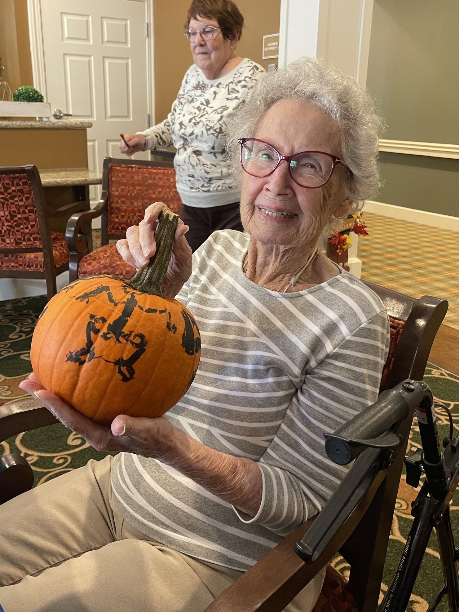 "Woman with glasses smiling, holding a pumpkin with a painted face, seated indoors.